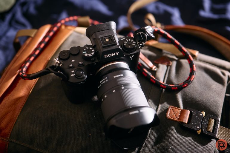 A Sony camera with a large lens rests on a canvas and leather bag in natural light.