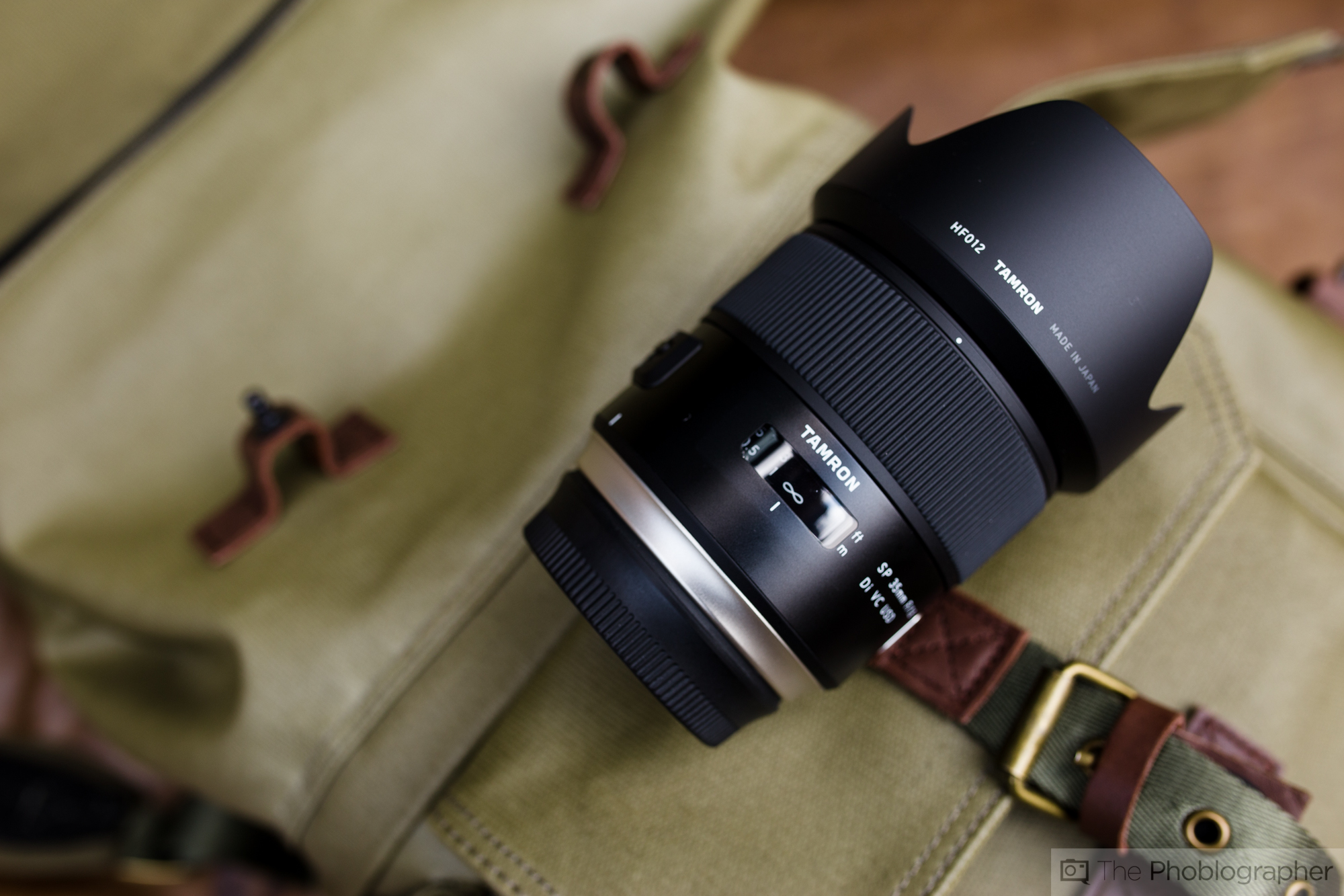 A Tamron camera lens rests on a khaki backpack with leather straps and brass buckle.