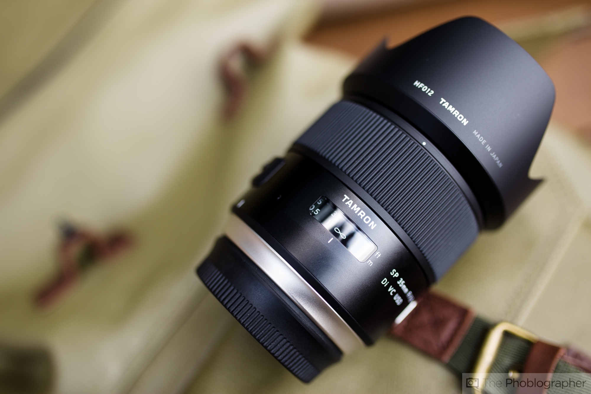 A Tamron camera lens rests on a fabric surface with a brown strap in the background.