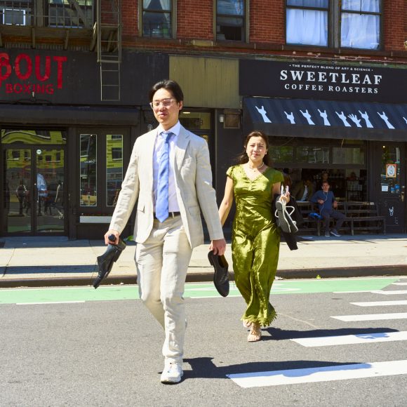 A man in a suit and a woman in a green dress walk barefoot across a city street, smiling.
