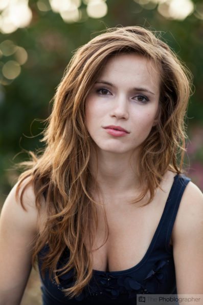 A young woman with long brown hair looks at the camera outdoors, wearing a navy blue top.