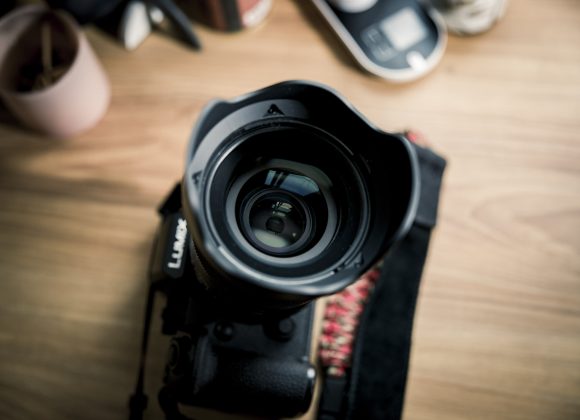 A close-up of a camera lens on a wooden desk with blurred objects in the background.