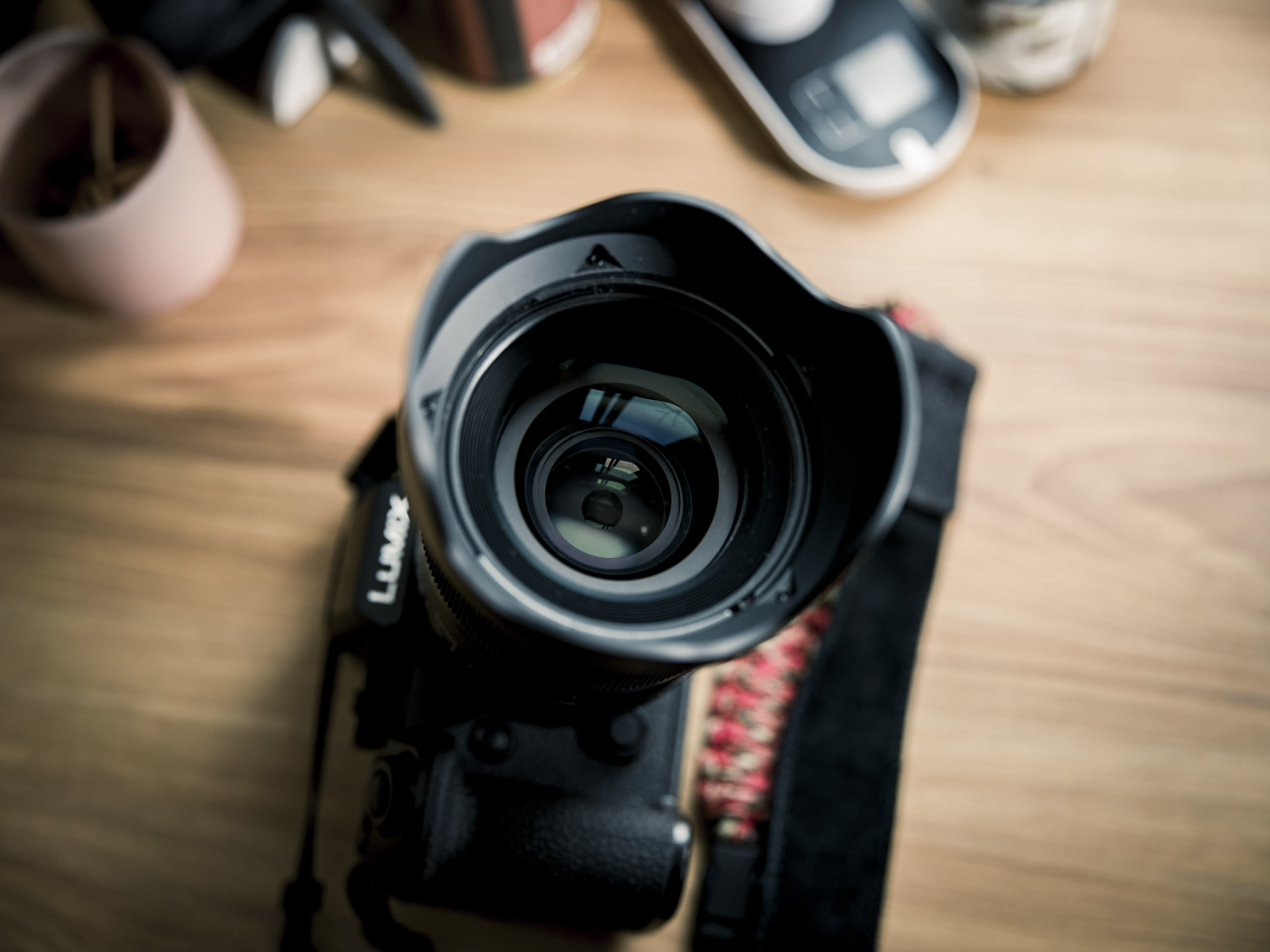 A close-up of a camera lens on a wooden desk with blurred objects in the background.