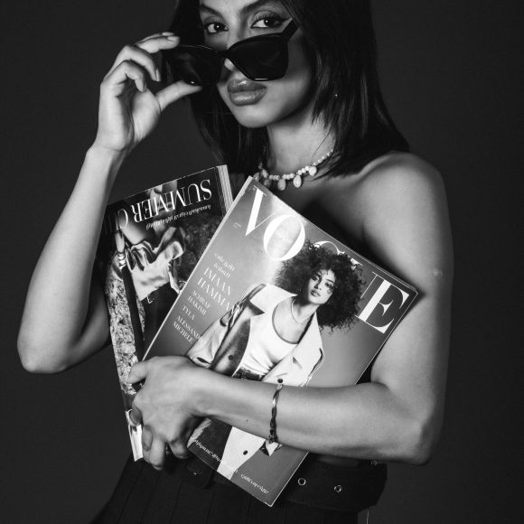 Woman in black dress holds Vogue and Another magazines, lifting sunglasses, with a serious expression. Black and white.