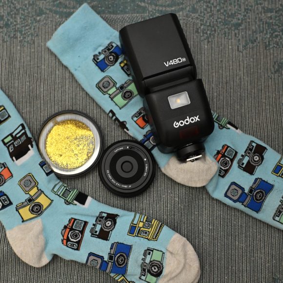 Two blue socks with camera prints, a camera flash, a camera lens, and a tin with yellow glitter.