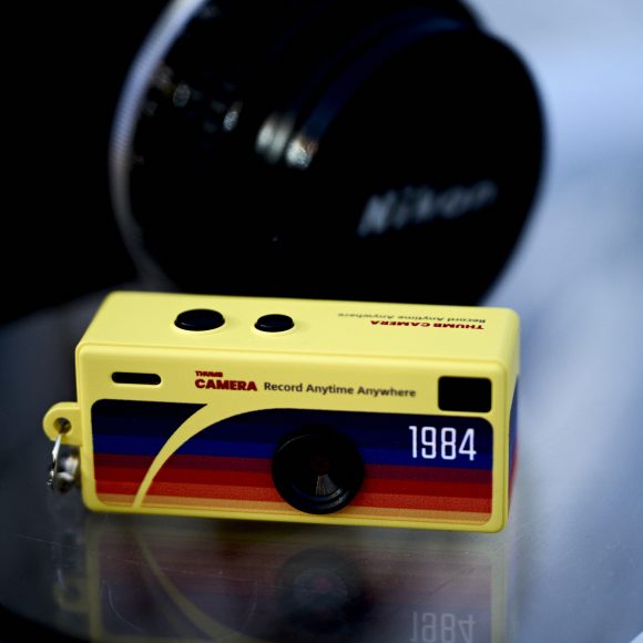 A small yellow toy camera keychain with 1984 and colorful stripes, next to a Nikon camera lens.