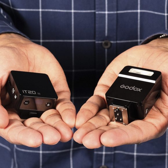 A person holds two small black camera accessories, one labeled IT20 and the other Godox, in their palms.