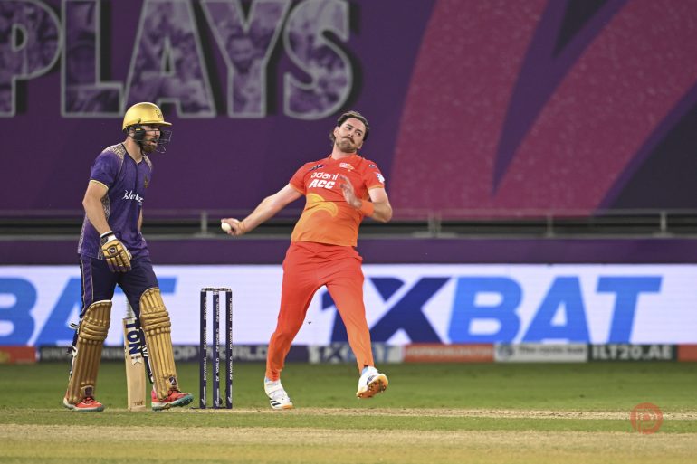A cricket bowler in orange delivers the ball as a batter in purple watches during a match.