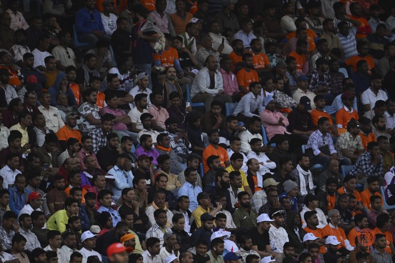 A large crowd of people, many in orange shirts and white caps, seated closely together in stadium stands.