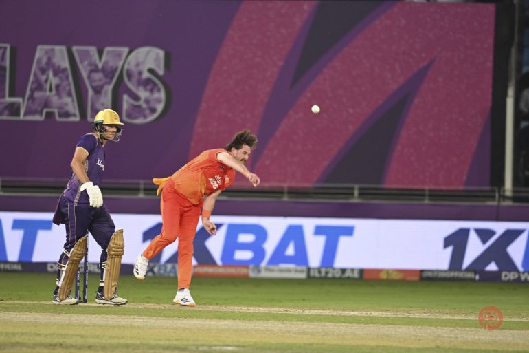 A cricket bowler in orange delivers the ball as a batter in purple stands ready at the wicket on the pitch.