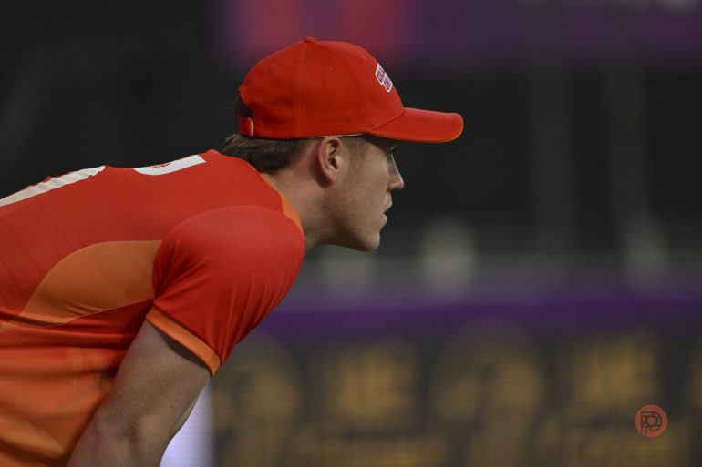A cricket player in an orange uniform and cap looks intently to the side during a game.