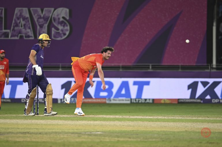 A cricket bowler in orange delivers the ball as a batsman in yellow and purple watches, on a stadium field.
