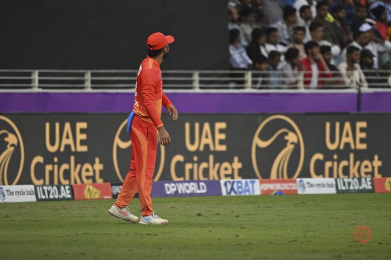 Cricket player in orange kit stands on field, with UAE Cricket ads and a crowd in the background.
