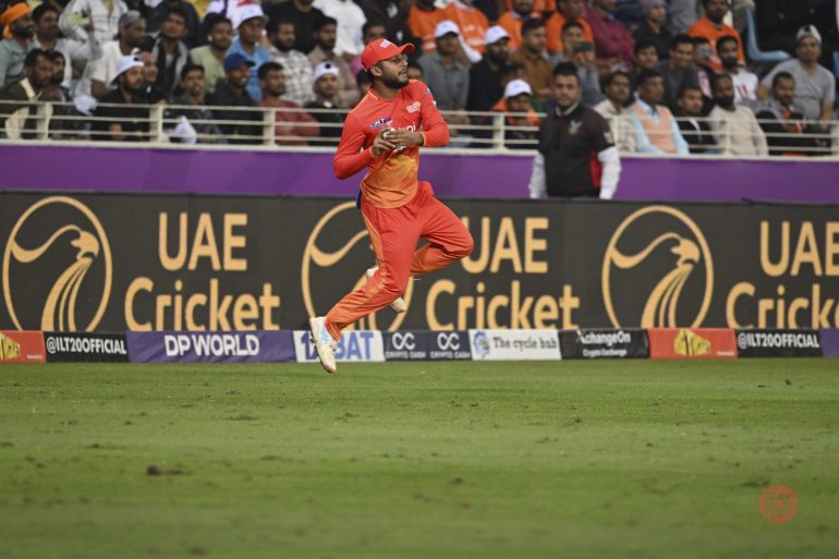 A cricket player in red uniform fields the ball during a match, with spectators in the background.
