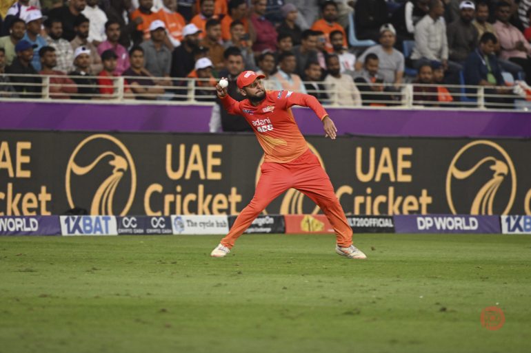 A cricketer in an orange uniform throws a ball on a field, with a crowd watching from the stands.