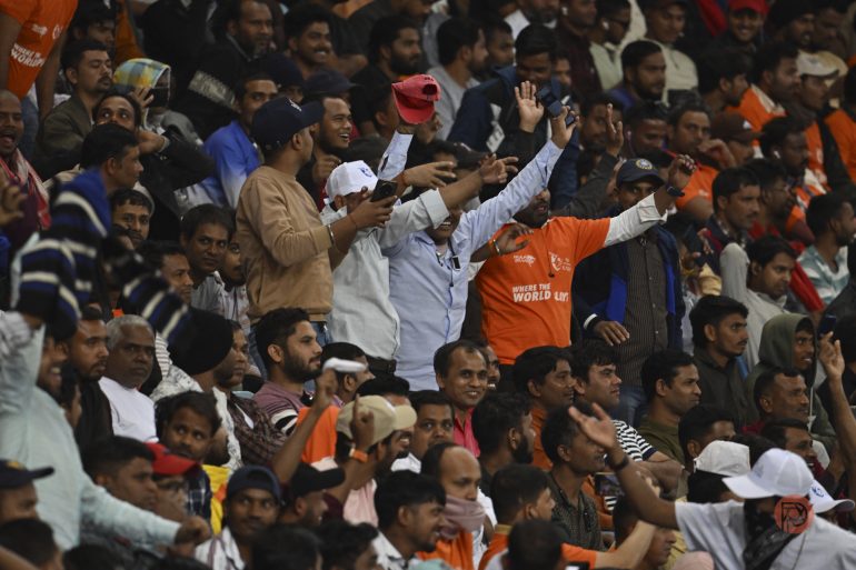 A large, enthusiastic crowd of cricket fans cheer and raise their hands in a stadium.