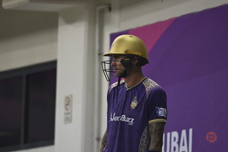 A cricket player in purple and gold gear stands indoors, ready to bat, with a purple wall behind him.