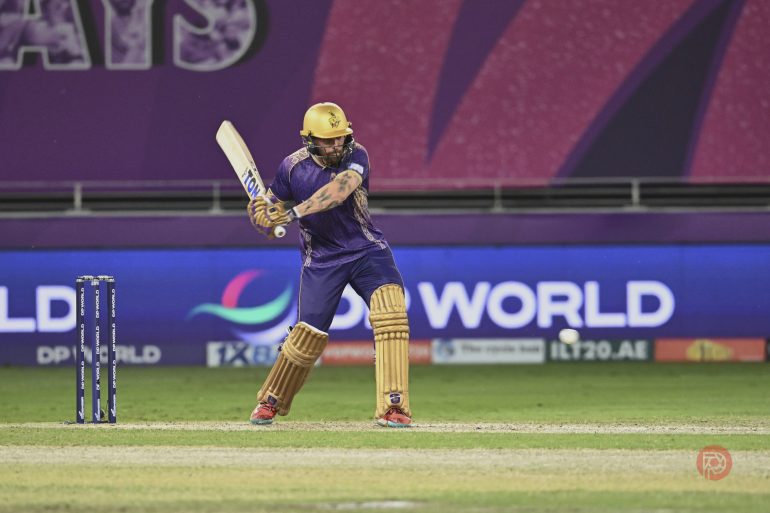 Cricketer in purple and gold uniform batting on a cricket field during a match.