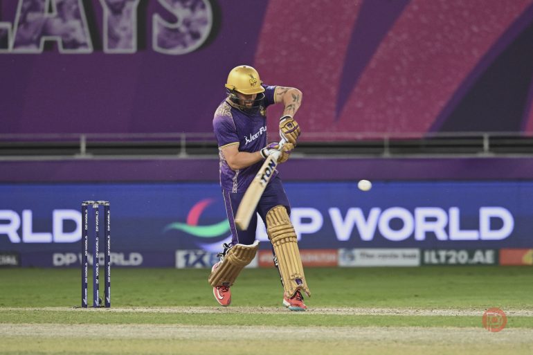 Cricketer in purple uniform playing a shot on a cricket field during a match.