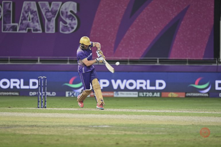 A cricket player in purple and yellow gear hits a ball on a stadium field during a match.