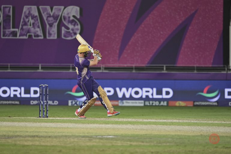 Cricket player in purple and gold uniform hitting a shot on a stadium field during a match.