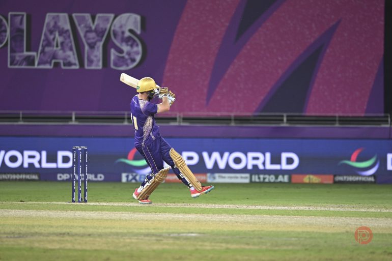 A cricket player in purple hits a shot on a field with advertising boards in the background.