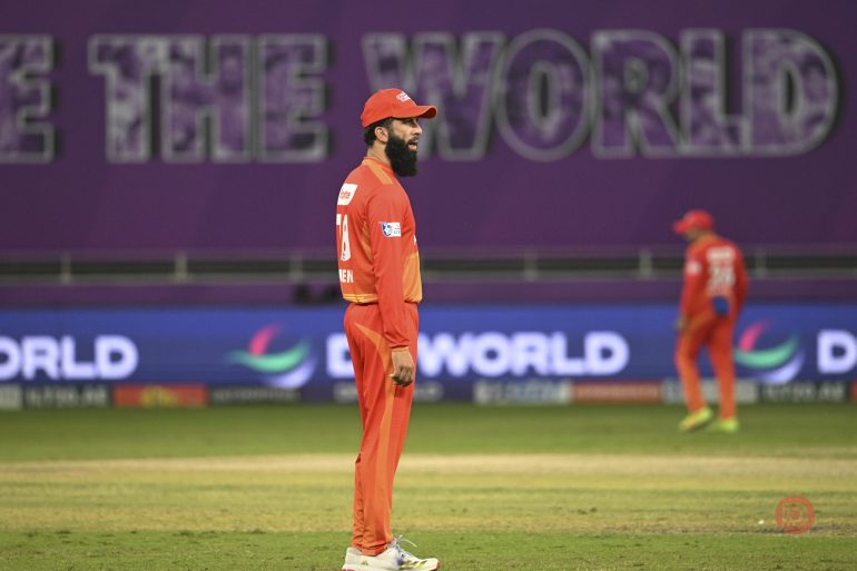 A cricket player in an orange uniform stands on the field, with another player and signage in the background.