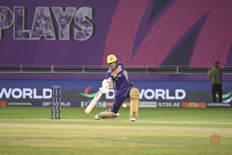 A cricket player in purple bats during a match, with stumps and advertising boards in the background.