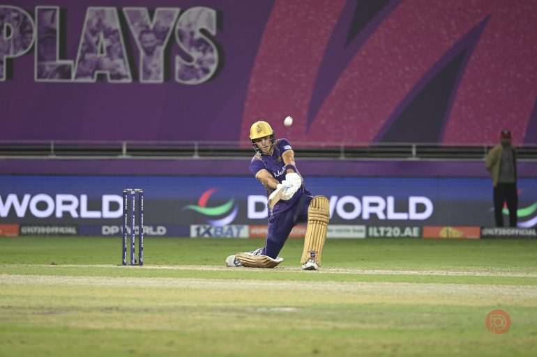 A cricket player in purple uniform plays a shot on a cricket field during a match.