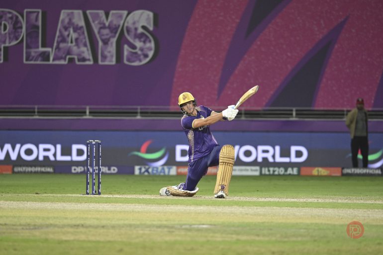 A cricketer in purple uniform plays a shot while kneeling on one knee during a match.