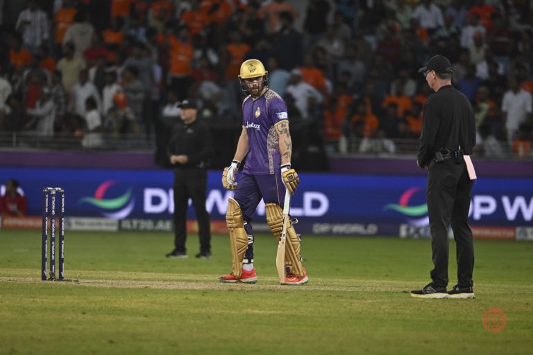 A cricket player in purple walks off the field with his bat while an umpire stands nearby; crowd in background.