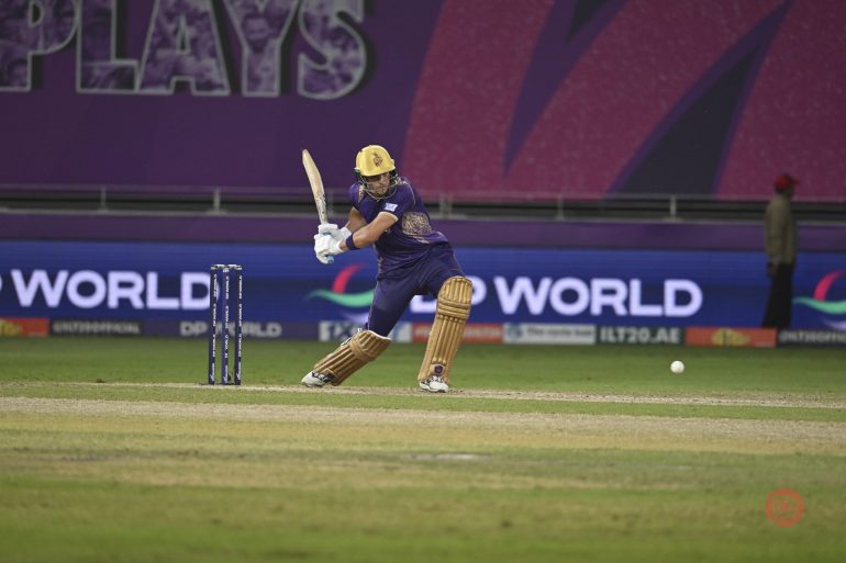 A cricketer in purple and yellow gear plays a shot during a cricket match on a green field.