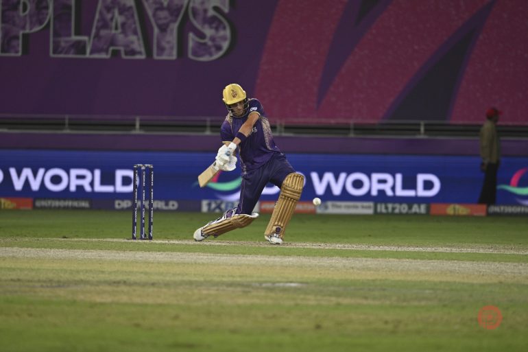 A cricket player in purple bats during a match on a lit stadium field with a purple backdrop.