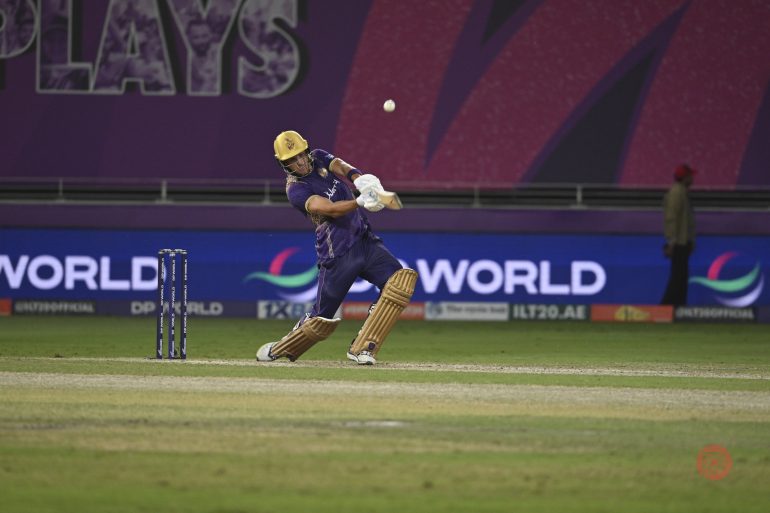 A cricket player in purple hits the ball on a stadium field during a match.