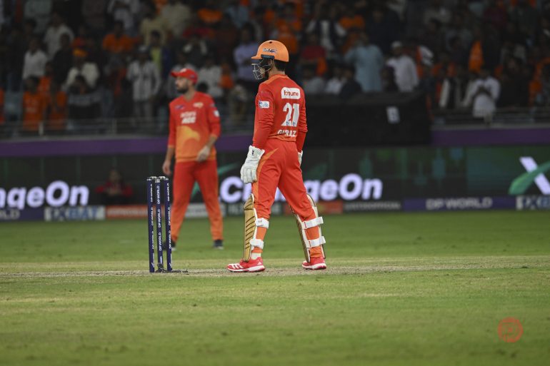 Cricket players in orange uniforms on the field during a match, with stumps and crowd in the background.