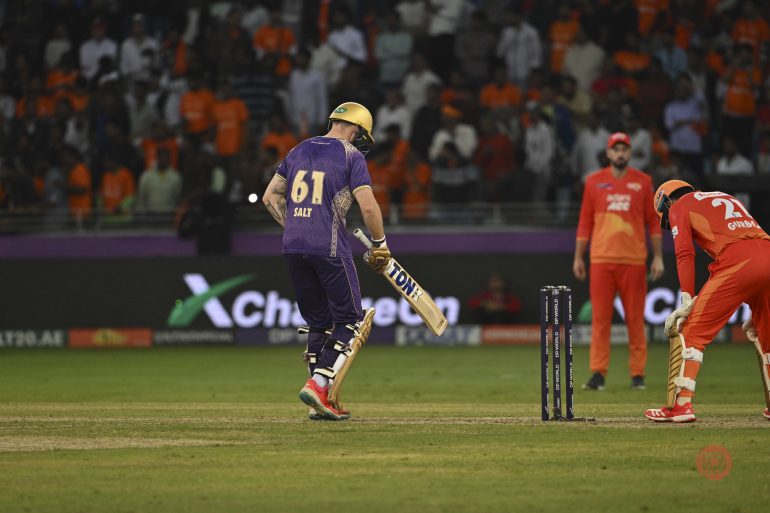 A cricket batter in purple jersey walks away after being bowled out, with fielders in orange in the background.