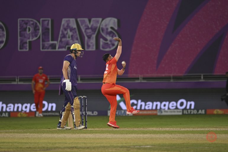 A cricket bowler delivers the ball as the batsman waits, during a night match on a brightly lit field.