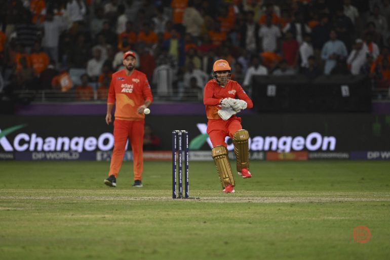A cricket wicketkeeper in orange gear prepares to catch the ball, with another player and crowd in the background.