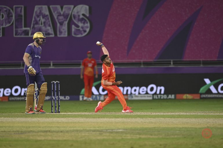A cricket bowler in orange delivers a ball as a batter in purple waits at the crease.