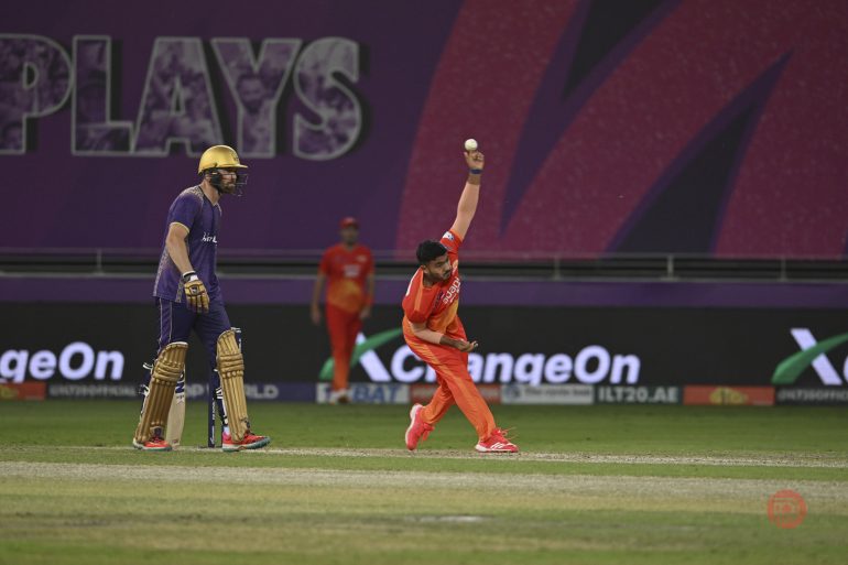 A cricket bowler in orange delivers a ball as a batter in purple watches, under stadium lights.