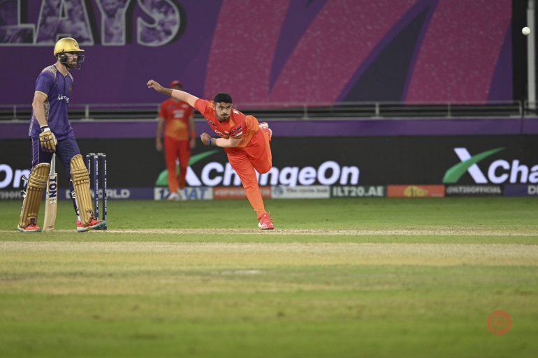 A cricket bowler in orange delivers the ball as a batsman in purple waits at the crease during a match.