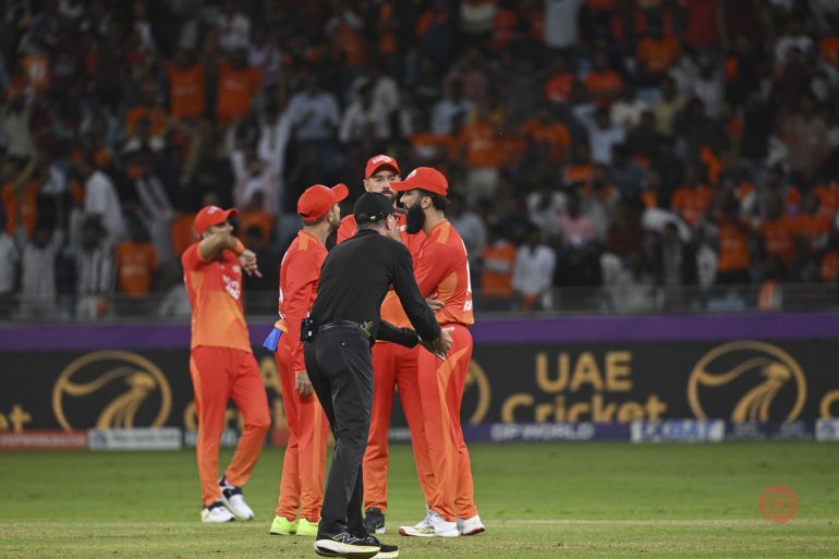 Cricket players in orange uniforms celebrate on field with umpire; crowd visible in the background.
