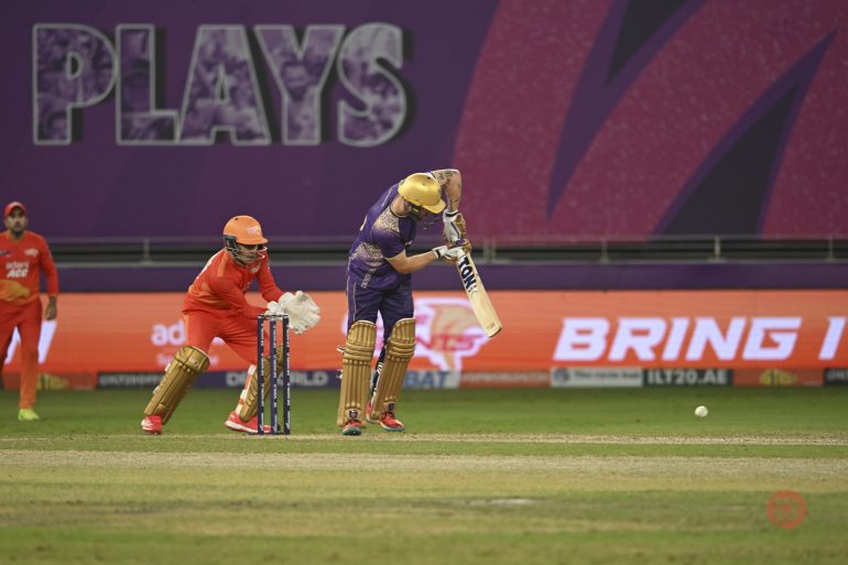 A cricket batsman in purple plays a shot as the wicketkeeper in orange prepares to catch the ball.