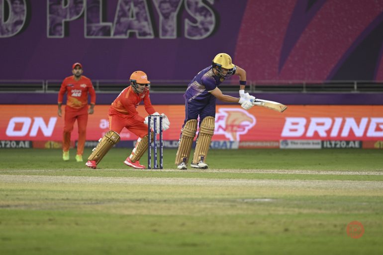 A cricket batsman in purple plays a shot as the wicketkeeper and fielder in orange watch closely.