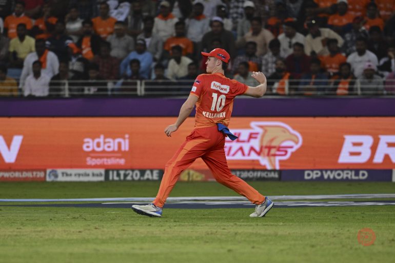 A cricket player in an orange uniform throws a ball on the field with a crowd watching in the background.