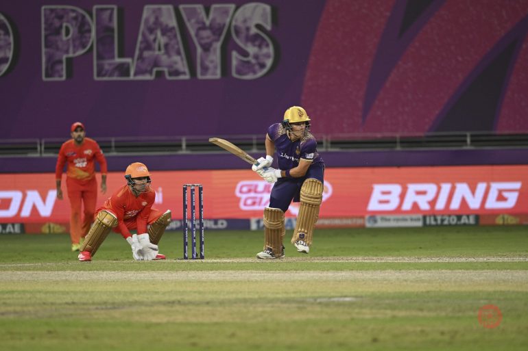 A cricket batter plays a shot as the wicketkeeper and fielders watch during a match in a stadium.