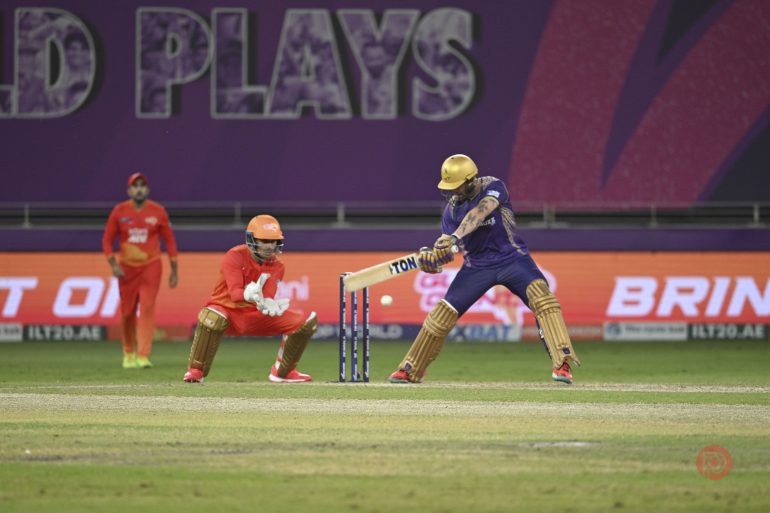 A cricket batsman in purple plays a shot as the wicketkeeper in orange crouches behind the stumps.