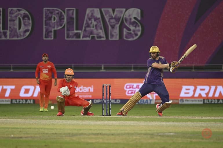 A cricket batsman plays a shot as the wicketkeeper and a fielder watch during a match.