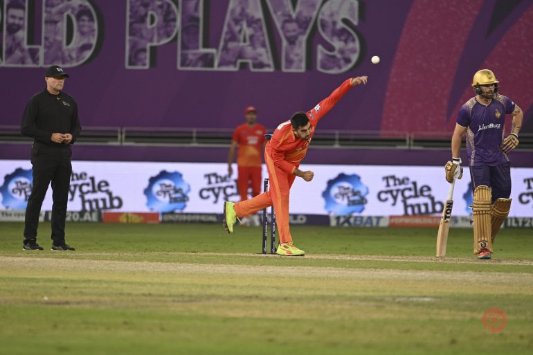A cricket bowler in orange delivers the ball as the batsman in purple and umpire watch on the field.