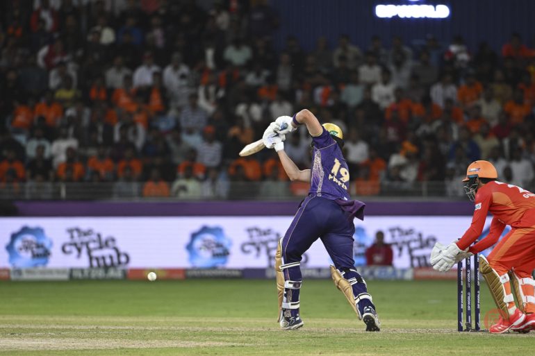 A cricket batsman in purple hits the ball as a wicketkeeper in orange crouches behind the stumps.